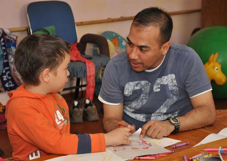 A father interacting with his son who is drawing a picture. He could be portraying the style of teacher- counselor or athletic coach.